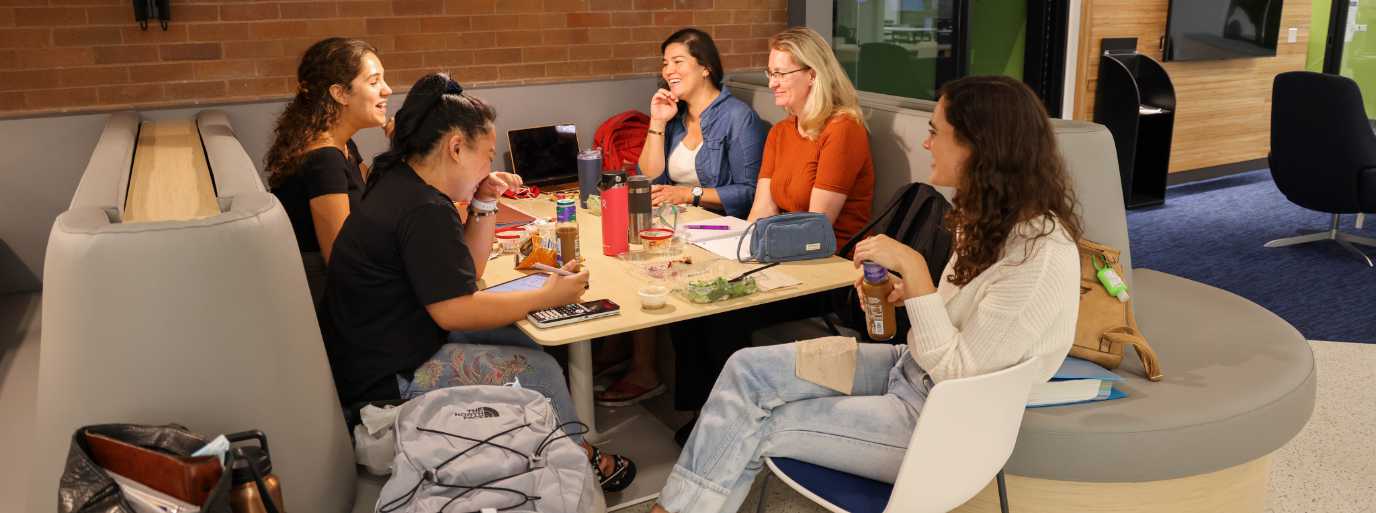 A group of female students sit around a table in the cafeteria smiling and laughing.