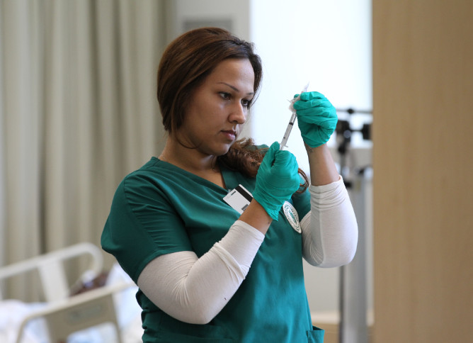 An Oakton nursing student wearing green scrubs checks the fluid level on a needle. 