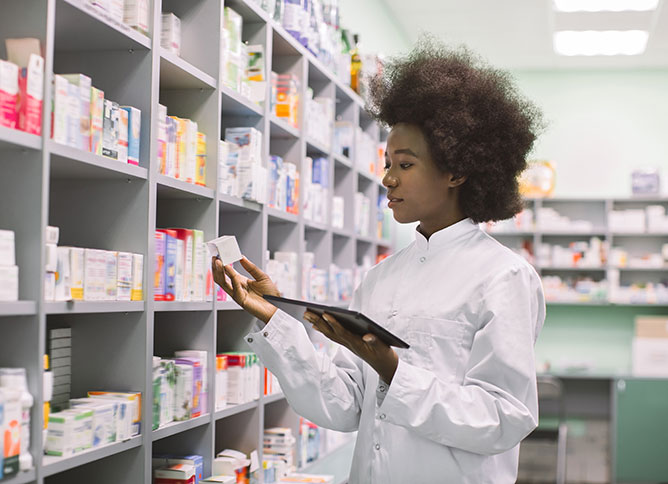 An African-American Pharmacy Technician holds an iPad while looking at medication in what looks to be a pharmacy.