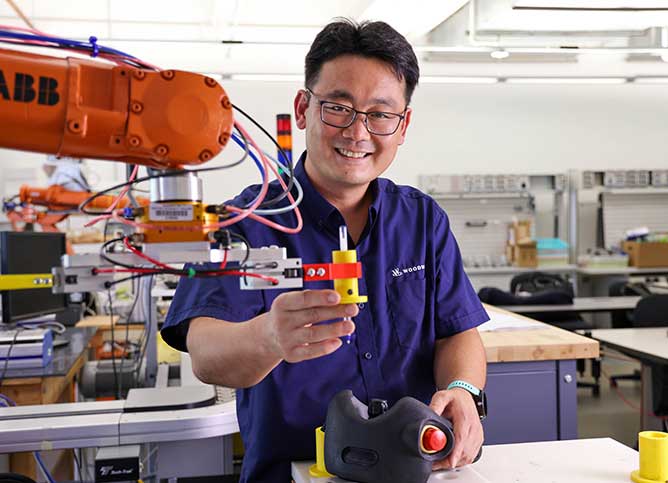 Yong Yoo wearing a blue shirt and glasses working in a classroom with manufacturing equipment