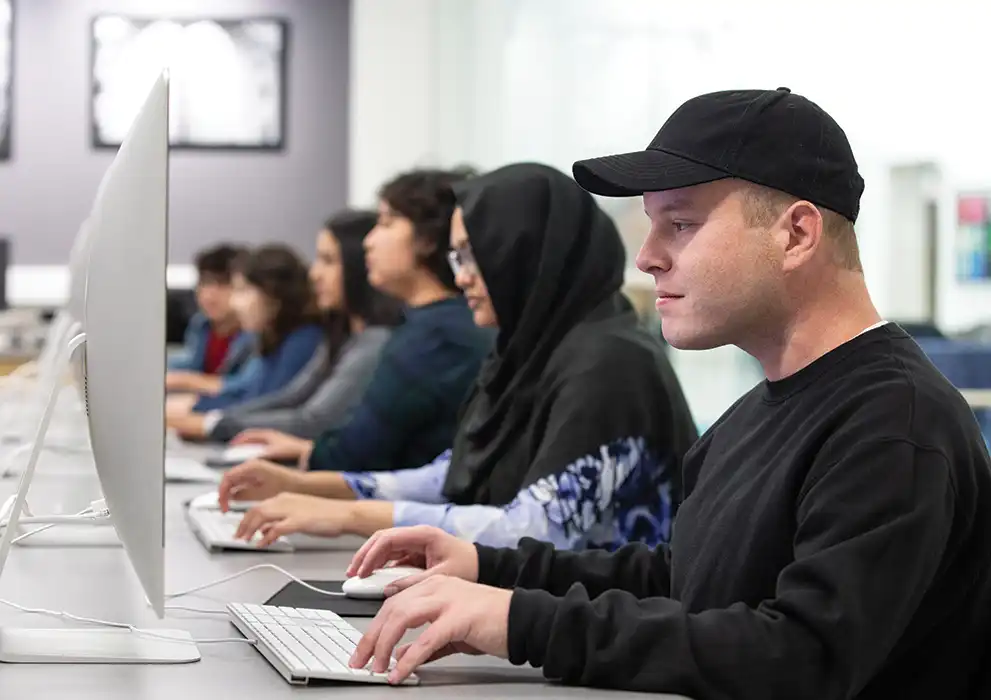 A student wearing a black hat and sweatshirt looks at a computer at Oakton College.