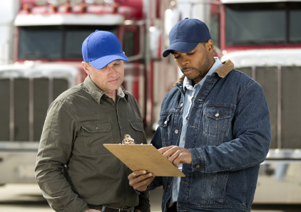 Two adults look at a clipboard while standing in front of two tractor-trailers.